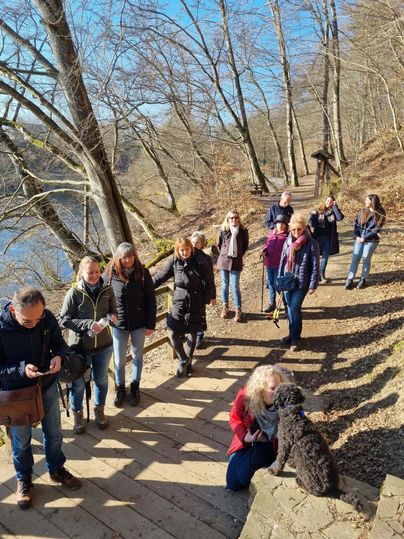 Eine Gruppe von Menschen wandert auf einem Holzweg entlang eines Sees. Einige stehen zusammen, während andere mit einem Hund interagieren.
