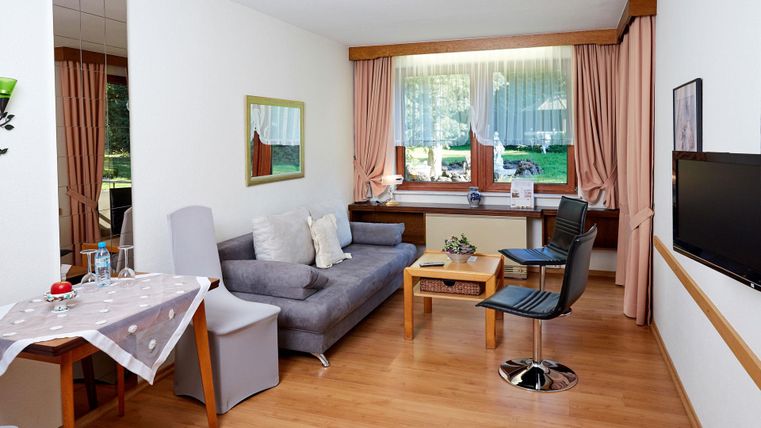 Ein gemütlicher Wohnbereich mit einem grauen Sofa, einem Tisch und einem Stuhl. Große Fenster sorgen für viel Tageslicht und einen Blick ins Freie.