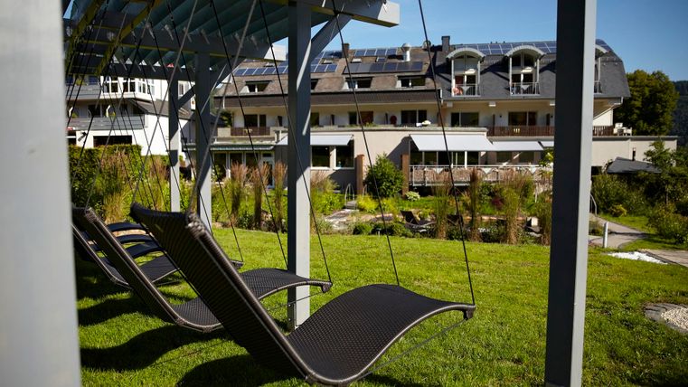 Eine gemütliche Liegefläche unter einem Vordach in einem schönen Garten. Im Hintergrund sind moderne Gebäude und Grünanlagen sichtbar.