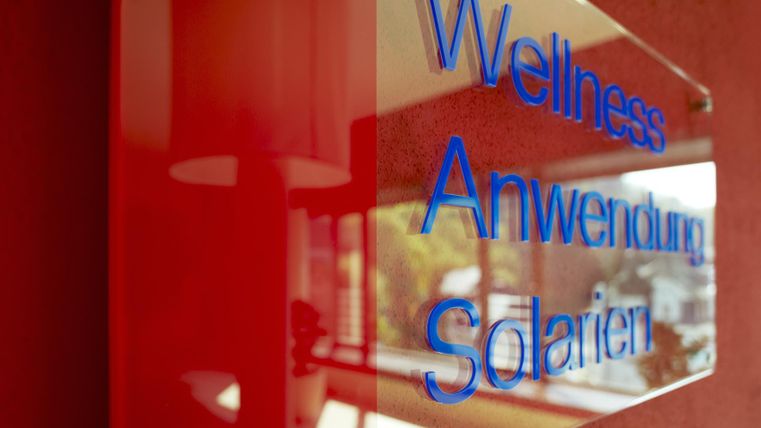 Ein Schild, das die Begriffe „Wellness“, „Anwendung“ und „Solarien“ zeigt. Der Hintergrund ist rot und das Schild hat einen modernen, eleganten Look.