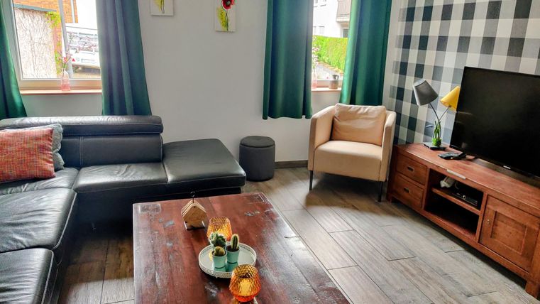 Ein modernes Wohnzimmer mit einem schwarzen Sofa und einem hellen Sessel. Ein Holzkaffee-Tisch steht im Zentrum, umgeben von dekorativen Elementen und einem Fernseher.