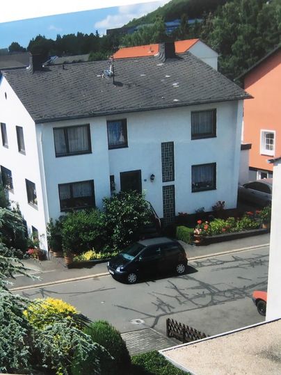 Ein weißes Wohnhaus mit Fensterläden und grünem Vorgarten. Auf der Straße parkt ein schwarzes Auto.