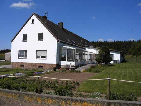 Ein großes, weißes Haus mit einem Wintergarten und einer gepflegten Rasenfläche. Im Hintergrund sind grüne Bäume und ein klarer Himmel zu sehen.