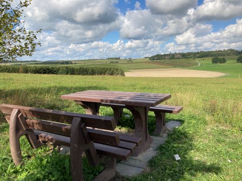 Holztisch und Bänke auf einer Wiese mit weitem Blick über Felder und Hügel unter einem bewölkten Himmel.