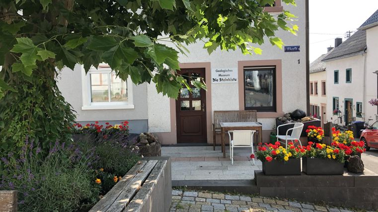 Ein freundliches Haus mit einem einladenden Eingangsbereich. Vor dem Haus stehen bunte Blumen und es gibt eine kleine Sitzgelegenheit.