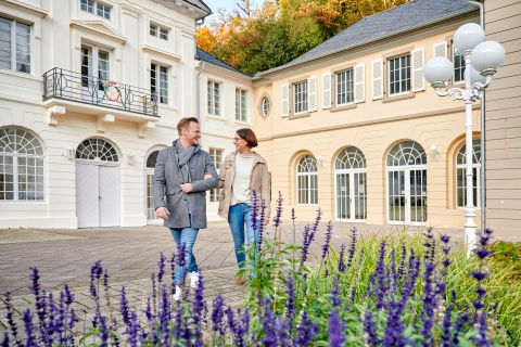 Ein Paar spaziert durch einen malerischen Garten mit lila Blumen. Im Hintergrund sind elegante Gebäude mit großen Fenstern zu sehen.