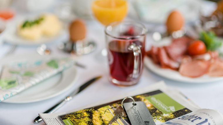 Ein reichhaltiges Frühstücksbuffet mit verschiedenen Speisen, darunter Eier, Wurst und frischer Saft. Auf dem Tisch liegen auch eine Zeitung und ein Schlüsselanhänger.