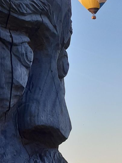 Eine große Holzskulptur eines Gesichts steht im Vordergrund. Im Hintergrund schwebt ein Heißluftballon mit dem Schriftzug „Technis“.