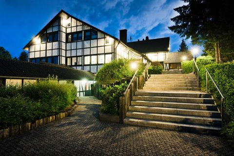 Ein modernes Gebäude mit großen Fenstern, beleuchtet bei Nacht. Eine steinerne Treppe führt zu dem Eingang, umgeben von grünen Büschen.