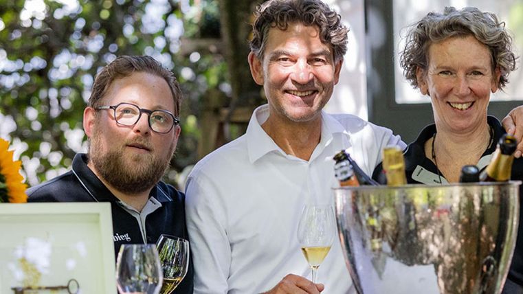 Drei Personen lächeln begeistert vor einer Flasche Sekt und Gläsern. Im Hintergrund sind grüne Bäume und eine idyllische Szene zu sehen.