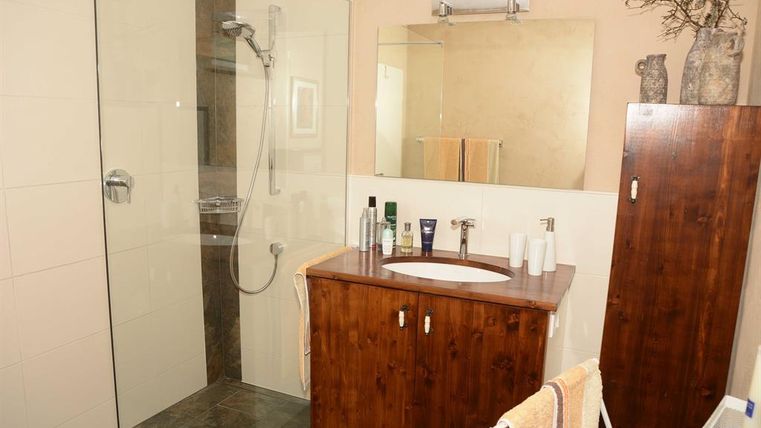 Ein modernes Badezimmer mit einer Duschkabine aus Glas und einem Holzmöbel. Der Spiegel reflektiert wenig Licht, und verschiedene Pflegeprodukte sind auf dem Waschtisch platziert.