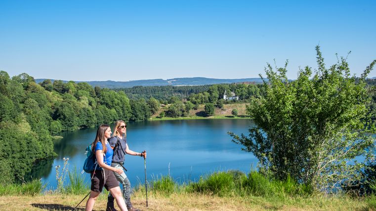 Zwei Wanderer gehen auf einem Hügel entlang eines kleinen Sees. Im Hintergrund befindet sich eine hügelige, waldige Landschaft, aus welcher eine kleine Kapelle herausragt.