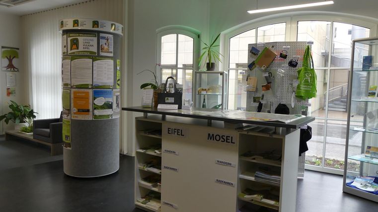 Ein moderner Empfangsbereich mit einem hohen Tisch und Informationsständen. Das Licht kommt durch große Fenster und die Umgebung wirkt einladend und freundlich.