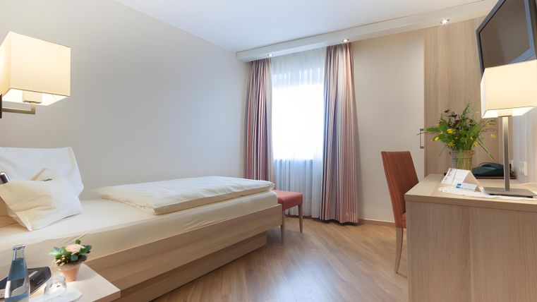 Ein gemütliches Hotelzimmer mit einem Einzelbett und einem Schreibtisch. Die Wände sind hell, und es gibt Fenster mit Vorhängen sowie einen Fernseher.