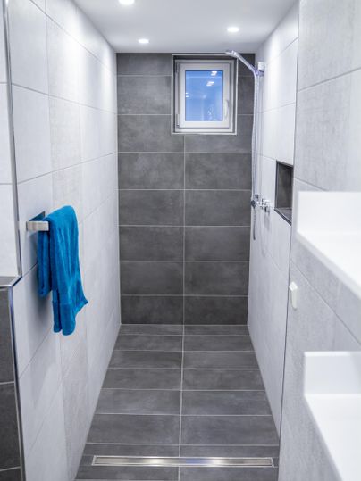 Eine moderne Dusche mit grauen Fliesen und hellem Licht. Ein blauer Handtuch hängt an der Wand.