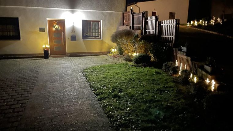 Eine gemütliche Einfahrt zu einem Haus bei Nacht, beleuchtet von warmen Lichtern. Der Rasen ist gepflegt und es gibt einen dekorativen Zaun.
