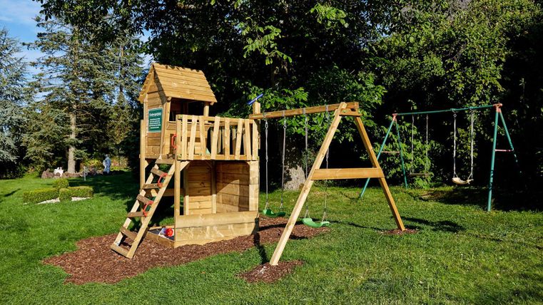 Ein Spielplatz mit einer Holzhütte und einer Sch swinging. Die Anlage befindet sich auf einer grünen Wiese umgeben von Bäumen.