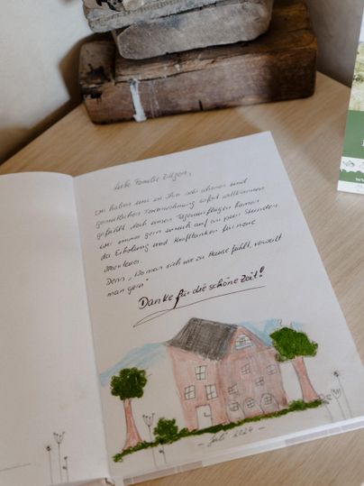 An open book with a handwritten note and a drawing of a house surrounded by trees. It is sitting on a table next to a wooden block and other decorative objects.