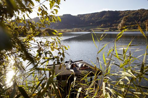 Ein ruhiger See umgeben von Ufervegetation und sanften Hügeln. Auf einem Holzsteg entspannen zwei Personen in der Sonne.