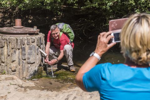 A man is filling a bottle with water from a spring. A woman is happily taking a picture of him from behind.