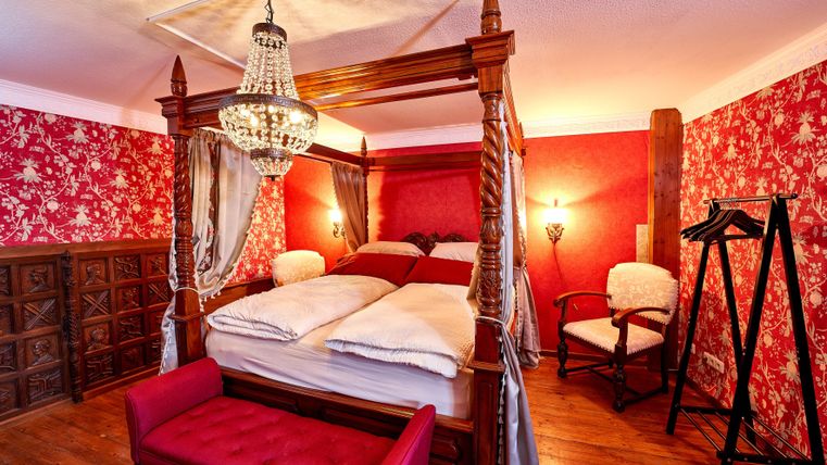 Ein elegantes Schlafzimmer mit einem Himmelbett und roter Tapete. Der Raum ist gemütlich eingerichtet und strahlt eine warme Atmosphäre aus.
