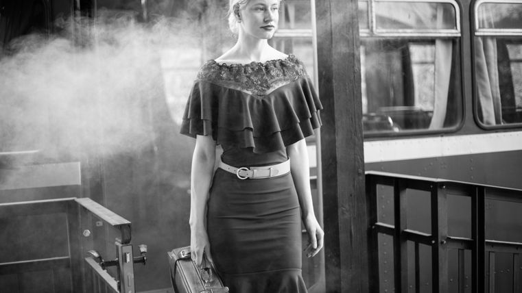 Eine elegante Frau mit einer Aktentasche steht in einem retro-inspirierten Bahnhof. Sie trägt ein schickes, finsteres Outfit und wirkt selbstbewusst.
