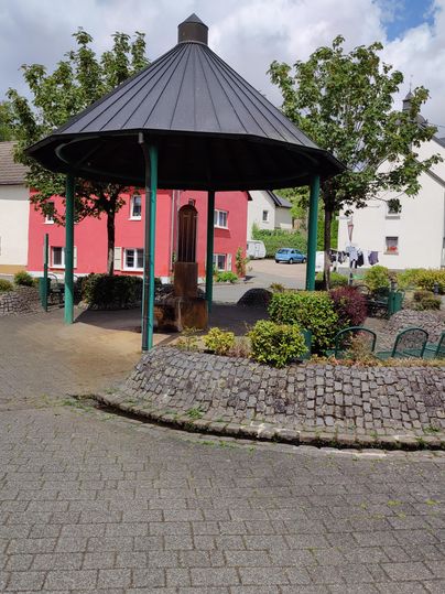 Ein Pavillon in einem kleinen Park, umgeben von Bäumen und Sträuchern. Im Hintergrund sind farbige Häuser zu sehen.