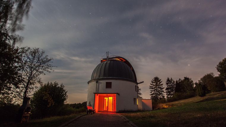 Eine Astronomieteleskop observatorium bei Nacht mit einem leuchtend roten Eingang. Umgeben von Bäumen und einem klaren Himmel.