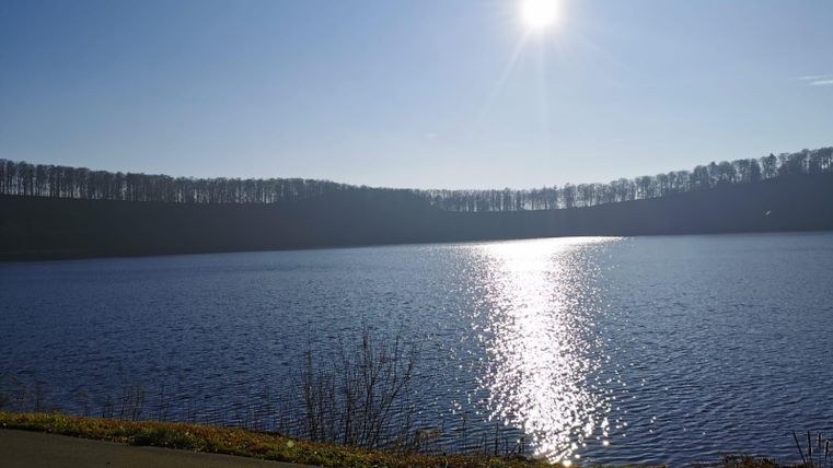 Ein ruhiger See mit spiegelndem Wasser und einer sonnigen Atmosphäre. Im Hintergrund sind bewaldete Hügel zu sehen.