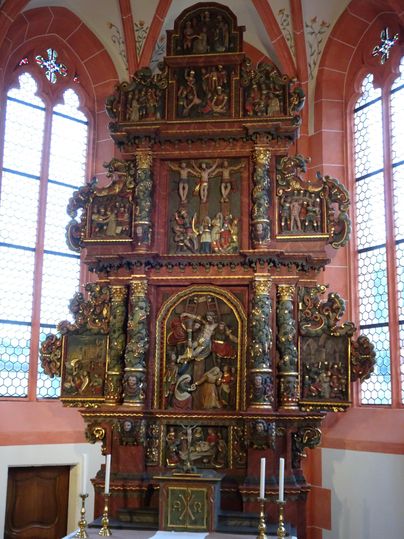 Ein kunstvoll geschnitzter Altar aus Holz, verziert mit religiösen Figuren. Der Altar steht in einer Kirche und hat große Fenster, die Licht hineinlassen.