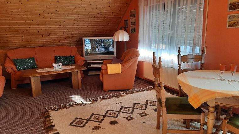 Ein gemütliches Wohnzimmer mit orangen Wänden und einem Holzboden. Die Einrichtung umfasst eine Sitzecke mit Couches, einen Tisch und einen Fernseher.