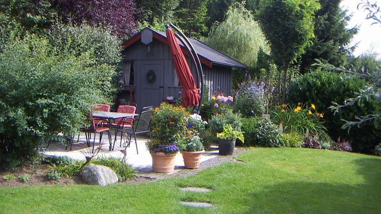Ein hübscher Garten mit bunten Blumen und einem gemütlichen Sitzbereich. Im Hintergrund steht ein kleines Gartenhaus umgeben von Bäumen.