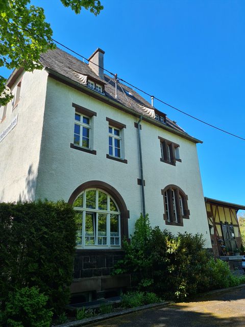 Ein schönes, mehrstöckiges Haus mit weißen Wänden und mehreren Fenstern. Umgeben von kurzen Sträuchern und Bäumen unter einem klaren blauen Himmel.