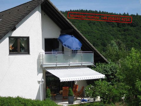 Ein helles Haus mit einem Balkon, auf dem ein blauer Sonnenschirm steht. Im Hintergrund sind grüne Hügel zu sehen.