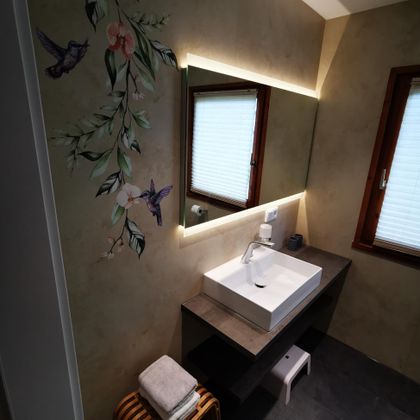 Ein modernes Badezimmer mit einem eleganten Waschbecken und einer großen Spiegelbeleuchtung. An der Wand gibt es eine dekorative Pflanze mit Vögeln.