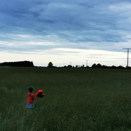 Ein Kind steht in einem Feld mit hohen Gräsern und hält einen roten Gegenstand. Der Himmel ist bewölkt und es gibt weitestgehend flache Landschaft.