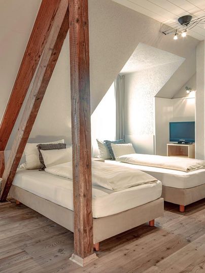 Ein gemütliches Schlafzimmer mit zwei Einzelbetten und Holzbalken. Natürliches Licht fällt durch das Fenster und es gibt einen kleinen Fernseher im Raum.