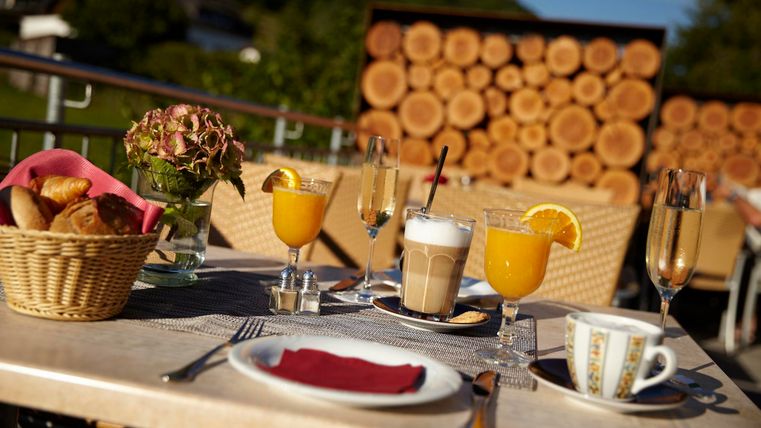 Ein gedeckter Tisch auf einer Terrasse mit frischem Gebäck, Säften und Kaffee. Im Hintergrund sind Gartenmöbel und Holzstämme sichtbar.