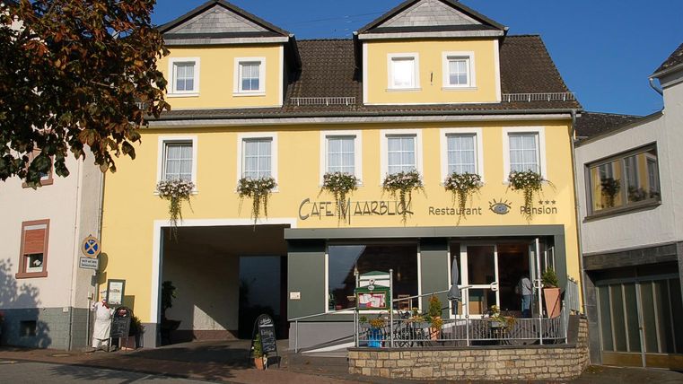 Ein gelbes Gebäude mit einem Café im Erdgeschoss. Vor den Fenstern hängen Blumenkästen, und die Fassade ist freundlich und einladend.