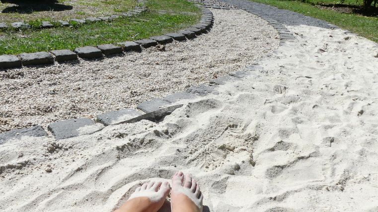 Zwei Füße im Sand auf einem gepflasterten Weg. Im Hintergrund sind gut gepflegte Grünflächen zu sehen.