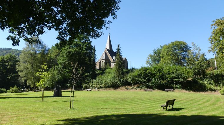 Eine schöne Wiese mit einem Blick auf eine Kirche und viel grünem Laub. Der Himmel ist blau und sonnig, ideal für einen entspannten Tag im Freien.