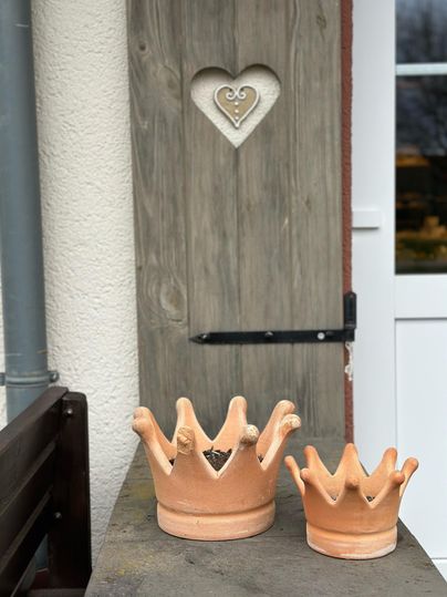 Zwei Tongefäße in Form von Kronen auf einer Holzoberfläche. Im Hintergrund ist eine Tür mit einem herzförmigen Ausschnitt sichtbar.