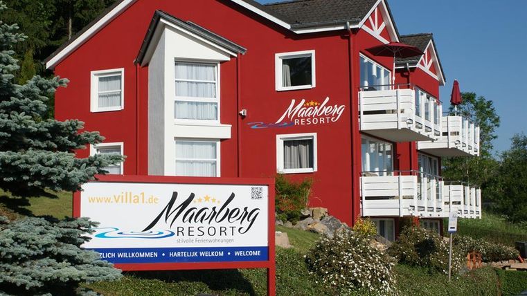 Ein rotes Gebäude mit dem Namen "Mahnberg Resort" und mehreren Balkonen. Ein Willkommensschild steht im Vordergrund, umgeben von grüner Landschaft.