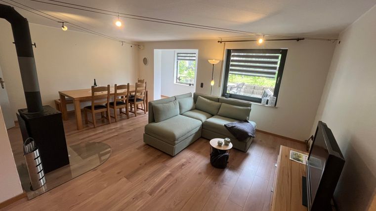 Ein helles Wohnzimmer mit einem bequemen Sofa und einem Kamin. Im Hintergrund steht ein Esstisch mit Stühlen und große Fenster bieten viel Licht.