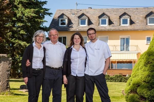 Eine vierköpfige Familie steht vor einem großen, gelben Haus mit einem gepflegten Garten. Alle Mitglieder lächeln und tragen helle Kleidung.