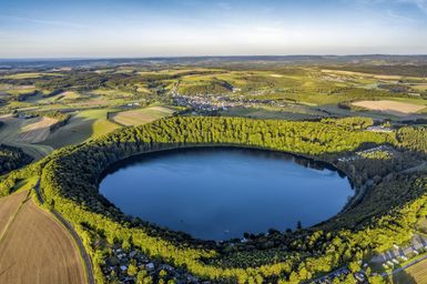 Ein schöner Kratersee, umgeben von grünen Wäldern und landwirtschaftlichen Feldern. Die Landschaft strahlt Ruhe und Naturverbundenheit aus.