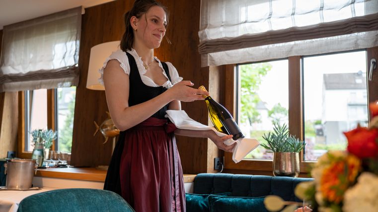 Eine Kellnerin in traditioneller Tracht hält eine Flasche Sekt und serviert sie. Im Hintergrund sind Fenster mit Blick ins Freie und eine stilvolle Einrichtung zu sehen.