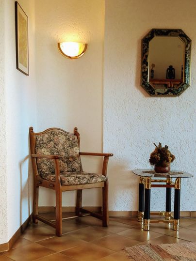 Ein gemütlicher Raum mit einem Stuhl, der mit Blumenmuster bezogen ist. Eine kleine Tisch mit einer Vase steht daneben, und ein Spiegel hängt an der Wand.