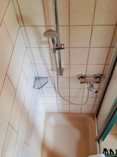Ein einfaches Badezimmer mit einer Dusche und Fliesen. Die Duscharmatur und eine Duschbrause sind sichtbar.