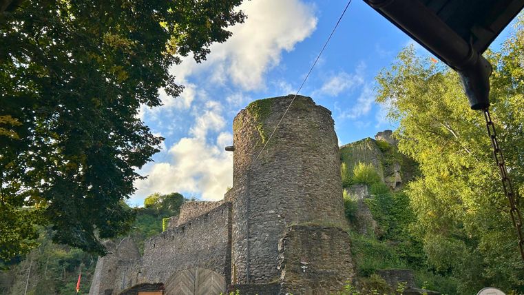 Eine alte Burgmauer umgeben von Bäumen und Hügeln. Der Himmel ist teilweise bewölkt und das Licht ist sanft.